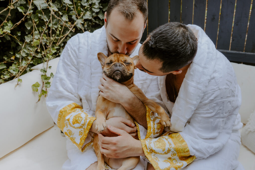 An lgbtqia+ couple in white robes lovingly holding their French Bulldog between them, both leaning in for a kiss on the dog's head, while preparing for the wedding day.